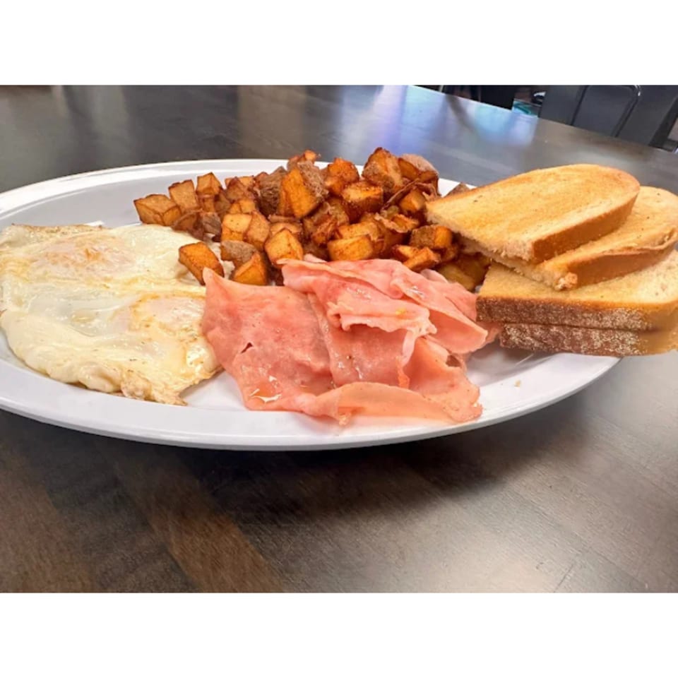 A deli breakfast plate with fried eggs, crispy home fries, sliced ham, and buttered toast.