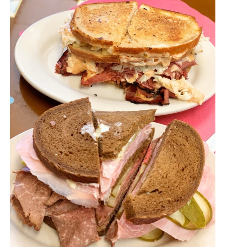 Two deli sandwiches on white plates, one a Reuben and the other a dark bread sandwich.